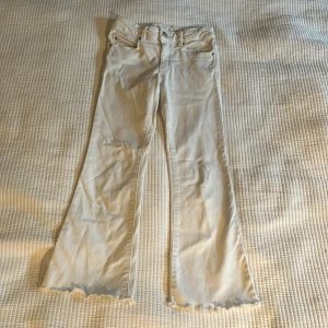 Vita bootcut jeans från Zara, stl 140 - Snygga vita jeans från Zara med bootcut passform och råa, fransiga benslut. Klassisk femficksmodell med knapp och dragkedja framtill. Perfekta för dig som vill ha en clean look med lite edge. Tillverkade i bomull för skön känsla.