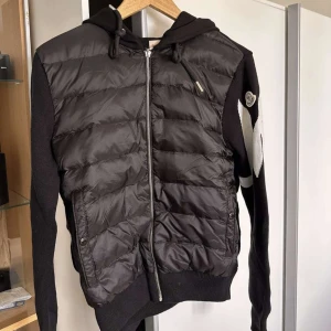 Moncler Logo-intarsia padded cardigan -  Säljer en helt ny och oanvänd Moncler Logo-Intarsia Padded Cardigan. Denna exklusiva hybridjacka i storlek S kombinerar en varm, dunjacka-inspirerad front med en elegant stickad rygg och ärmar. Jackan är prydd med en iögonfallande, stickad Moncler-logga på ena ärmen och det ikoniska Moncler-märket på den andra. Denna modell är perfekt för övergångssäsonger eller som ett stilfullt lager. Detaljer:  