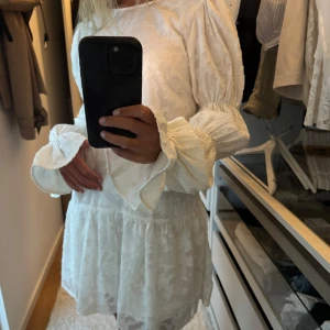 Vit kortklänning från gina - Superfin vit kortklänning med puffiga långa ärmar och volang vid ärmslut. Klänningen har strukturmönster i tyget, öppen rygg med knyt och en lätt volangkant nertill. Oanvänd, säljer då jag köpte för stor storlek❣️