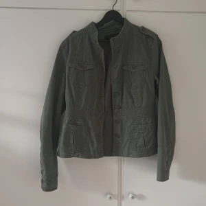 Grön militärinspirerad jacka från Vila - Snygg grön jacka från Vila med militärvibbar, ståkrage och knappar framtill. Jackan har fyra fickor med lock och axelklaffar som ger en cool look. Perfekt för lager-på-lager och passar till både jeans och kjol.