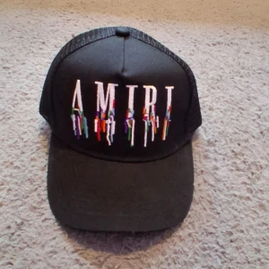 Svart keps från Amiri med broderi - Svart truckerkeps från Amiri med justerbar snapback och mesh på sidorna. Framtill finns ett stort broderat AMIRI-logo i vitt med färgglada detaljer som ger en cool drip-effekt. Perfekt för dig som vill sticka ut med streetstyle.
