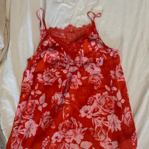 Röd blommig linnetopp från Gina Tricot - Supersöt röd linnetopp från Gina Tricot med rosa blommigt mönster och spetsdetaljer vid v-ringningen. Tunt och glansigt material med smala axelband, perfekt för en somrig look. Säljer då den inte kommer till användning!