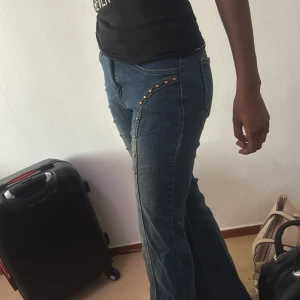 Blå bootcut jeans med nitar - Säljer ett par blå bootcut jeans med snygga nitar längs sidorna framtill. Jeansen har klassisk femficksdesign, normal midja och är tillverkade i denim. Perfekta för dig som vill sticka ut lite extra med detaljer och gillar en retrovibe.