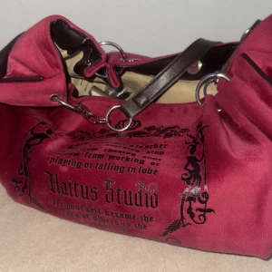 Röd axelväska Hiatus Studio i sammet - Cool röd axelväska från Hiatus Studio i mjuk sammet med svart tryck och dekorativa detaljer. Väskan har bruna axelremmar i skinnimitation, silverfärgade ringar och ett rymligt beige foder med innerfack och dragkedja. Perfekt för dig som gillar statement-accessoarer.