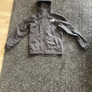 Grå vindjacka från The North Face - Snygg grå vindjacka från The North Face med huva och dragkedja framtill. Jackan har två sidofickor med dragkedja och en bröstficka, samt logga på bröstet och ryggen. Perfekt för blåsiga dagar och outdoor vibes.