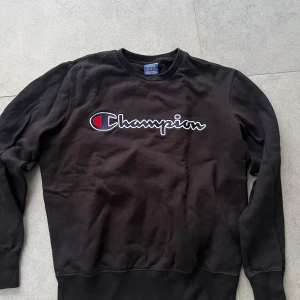 Svart Champion sweatshirt i bomull - Svart sweatshirt från Champion med stort broderat logotyptryck i vitt, blått och rött över bröstet. Klassisk rund halsringning och långa ärmar. Tillverkad i mjuk bomull, perfekt för en avslappnad och sportig stil.