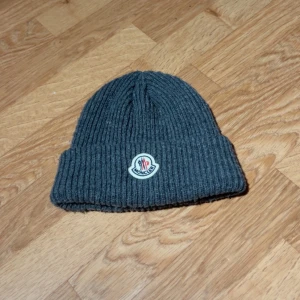 Grå ribbstickad mössa från Moncler - Snygg grå ribbstickad mössa från Moncler med klassisk uppvikt kant och broderad logotyp framtill. Perfekt för kalla dagar och ger en clean look. Tillverkad i mjukt ullmaterial som håller dig varm hela vintern.