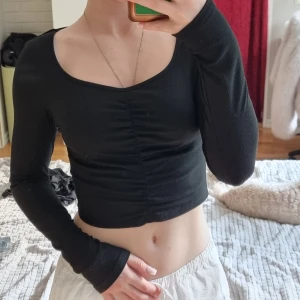 Svart croppad långärmad topp - Säljer en svart, croppad långärmad topp med snygg rynkdetalj framtill. Toppen har en djup, rundad urringning och är figurnära i modellen. Perfekt att styla med höga jeans eller kjol för en trendig look.