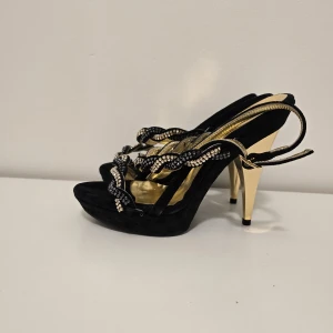 Svarta och guldiga pumps från Stefania Pellici - Eleganta pumps i svart mocka med höga guldklackar och glittrande detaljer. Skorna har vågformade remmar dekorerade med svarta och silvriga stenar samt öppen tå och guldfärgad innersula. Perfekta för dig som vill sticka ut.