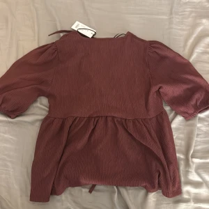 Vinröd blus med puffärm - Supersöt vinröd blus med puffiga trekvartsärmar och lätt rynkad nederdel. Blusen har en avslappnad passform och är gjord i ett mjukt, strukturerat tyg som ger en snygg look. Perfekt att styla med jeans eller kjol för en trendig vibe.