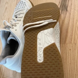 Vita Unihoc sneakers med grå detaljer - Snygga vita sneakers från Unihoc med grå detaljer och mönstrad ovandel. Skorna har snörning, platt sula i gummi och ventilerande meshpartier. Perfekta för inomhussport eller träning. Bekväm passform och sportig look.