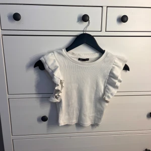 Vit volangtopp från Zara - Superfin vit topp från Zara med volangdetaljer på axlarna. Modellen är croppad och ärmlös, i ribbad bomull som känns mjuk mot huden. Perfekt för dig som gillar en söt och trendig look med det lilla extra.