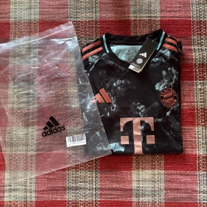 FC Bayern München Adidas fotbollströja S - Snygg FC Bayern München fotbollströja från Adidas i svart med unikt marmorerat mönster och detaljer i koppar/rosa. Tröjan har korta ärmar, klassiska Adidas-ränder på axlarna och klubbmärke på bröstet. 