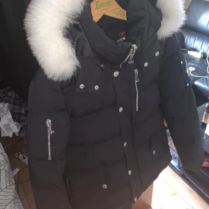 Svart Moose Knuckles dunjacka med päls - Säljer en svart dunjacka från Moose Knuckles med fluffig vit päls runt huvan. Jackan har flera silvriga dragkedjor och knappar, samt broderad logga på insidan. Perfekt för kalla dagar och riktigt bekväm. Storlek är m men funkar till L kvittot har tyvär tappats bort under en flytt för ett tag sedan. Hör gärna av er om ni vill ha mer information.söker endast seriösa köpare 