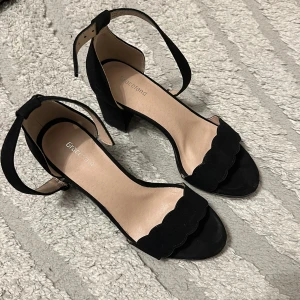 Svarta sandaler med klack från Graceland - Stilrena svarta sandaler från Graceland med öppen tå, blockklack och vågig kant framtill. Skorna har en justerbar rem runt ankeln och är tillverkade i mockaliknande material. Perfekta för dig som vill ha en chic och enkel look.