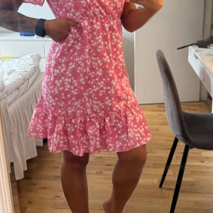 Rosa blommig kortklänning med volang - Supersöt rosa kortklänning med vita blommor, v-ringning och knappar framtill. Klänningen har korta ärmar, markerad midja och volangkant nertill. Perfekt för sommarens alla tillfällen!