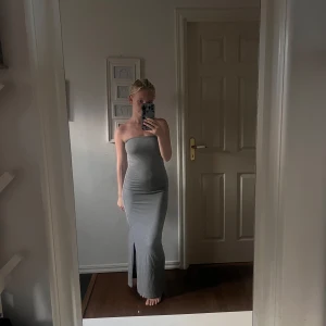 Grå axelbandslös långklänning - Säljer en stilren grå axelbandslös långklänning med tight passform och slits framtill. Klänningen är enkel men ändå snygg, perfekt om du gillar minimalistisk stil. Mjuk och stretchig, passar dig som vill ha något bekvämt men ändå trendigt.