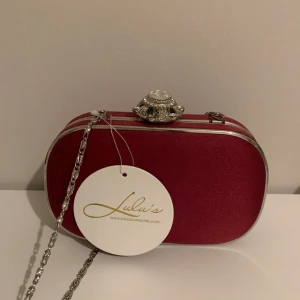 Röd kuvertväska från Lulus med kedja - Snygg röd kuvertväska från Lulus med silverfärgad kedja och en glittrig, stencool knäppning med stora stenar. Väskan har en hård, oval form och är klädd i ett sammetsliknande material. Perfekt för evenemang eller bal