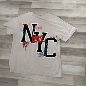 Beige NYC t-shirt med graffiti-tryck - Snygg beige t-shirt med stort NYC-tryck och färgglada graffiti-detaljer på ryggen. Framsidan har en mindre NYC-logga. T-shirten har klassisk passform, rund hals och är kortärmad. Perfekt för dig som gillar streetstyle och urban vibe. Over size T-shirt storlek S