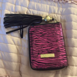 Zebramönstrad plånbok Steve Madden - Plånbok från Steve Madden i metallic rosa med svart zebramönster. Utsidan har en stor svart tofs med gulddetalj. Invändigt finns flera kortfack, myntficka med dragkedja och plats för ID. Materialet är syntet och ytan är glansig.