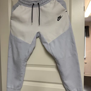 Ljusgrå/vita Nike Tech byxor - Nike Tech mjukisbyxor i ljusgrått och vitt med svart logga på framsidan. Byxorna har elastisk midja med snörning, muddar vid benslut och en svart dragkedja på bakfickan. Perfekta för chill eller träning.