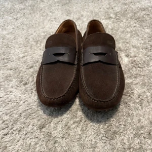 Bruna mockaloafers från Hugo Boss - Snygga loafers från Hugo Boss i mörkbrun mocka med klassisk penny strap i skinn över vristen. Skorna har rund tå och diskreta sömmar längs kanterna. Perfekta för en stilren och avslappnad look.