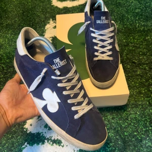 Blå sneakers från The Gallerist - Snygga blå sneakers från The Gallerist med vit klöverdetalj på sidan och vita snören. Ovandelen är i mocka och skon har en klassisk låg profil med vit och svart sula. Perfekta för dig som gillar stilrena men unika detaljer.