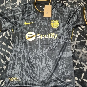 Barcelona svart fotbollströja Nike - Snygg svart Barcelona fotbollströja från Nike med gula detaljer och Spotify-logga på bröstet. Tröjan har korta ärmar och ett unikt grafiskt mönster inspirerat av stadens arkitektur. Perfekt för dig som vill sticka ut på planen eller läktaren.