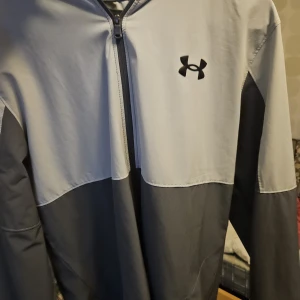 Grå vindjacka från Under Armour - Snygg tvåfärgad vindjacka från Under Armour i ljusgrått och mörkgrått. Jackan har huva, dragkedja framtill och logga på bröstet. Perfekt för träning eller när du vill ha en sportig look. Långärmad och med praktiska fickor.