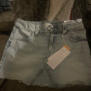 Ljusblå jeanskjol från Gina Tricot Young - Snygg ljusblå jeanskjol med låg midja från Gina Tricot Young. Kjolen har coola slitningar, rå kant nertill och broderade bakfickor med Y-detalj. Klassisk femficksmodell och bälteshällor, perfekt för en trendig vardagslook.