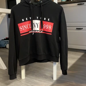 Svart hoodie från Divided med tryck - Svart hoodie från Divided med stort New York-tryck framtill i rött, vitt och svart. Tröjan har huva med snörning, långa ärmar och en croppad passform. Perfekt för dig som gillar streetstyle och vill ha en chill look.