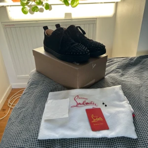 Christian Louboutin svarta sneakers med nitar - Svarta sneakers i mocka från Christian Louboutin med ikoniska röda sulor och svarta nitar på tån. 