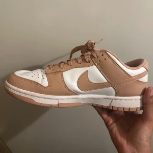 Nike Dunk Low i beige och vitt - Nike Dunk Low sneakers i en clean beige och vit färgkombination. Skon har klassisk låg siluett, snörning och rund tå. Ovandelen är i skinn med perforerade detaljer på tån för extra ventilation. Perfekt för dig som vill ha en stilren och trendig sneaker.