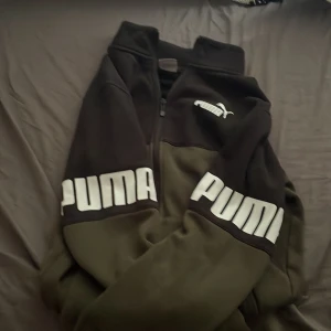 Svart och olivgrön jacka från Puma - Snygg svart och olivgrön jacka från Puma med dragkedja framtill och stora vita logotyper på ärmarna. Jackan har hög krage, två fickor och är tillverkad i mjukt syntetmaterial. Perfekt för dig som gillar sportig och street-inspirerad stil.