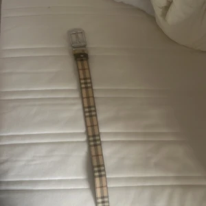 Rutig Burberry bälte i skinn - Snyggt bälte från Burberry med klassiskt rutigt mönster i beige, svart och rött. Bältet har en silverfärgad metallspänne och är gjort i skinn. Perfekt accessoar för att lyfta din outfit med en ikonisk touch.
