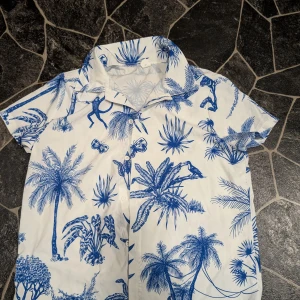 Vit kortärmad skjorta med blått djungelmönster stl 11-12 år  - Snygg vit skjorta med korta ärmar och blått djungelinspirerat mönster med apor, fjärilar, fåglar och palmer. Skjortan har klassisk krage och knäppning framtill. Perfekt för dig som vill sticka ut med en tropisk vibe.