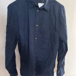 Mörkblå skjorta från H&M i bomull - Snygg mörkblå skjorta från H&M i storlek 170. Skjortan är tillverkad i bomull, har klassisk krage, bröstficka och knäppning framtill. Perfekt för dig som gillar en stilren och enkel look. Linne skjorta