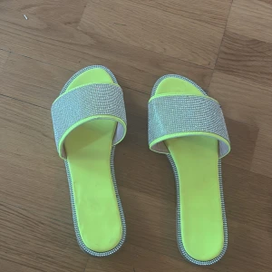 Neongula sandaler med strass - Snygga sandaler i neon-gult med breda band täckta av glittrande strass. Platt sula och fyrkantig tå ger en trendig vibe. Perfekta för att sticka ut i sommar! Kantade med silverfärgade detaljer för extra bling.