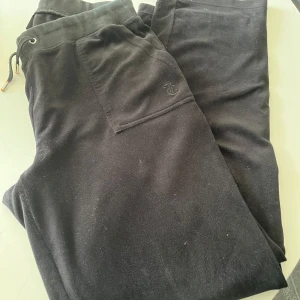 Svarta mjukisbyxor Juicy Couture L - Svarta mjukisbyxor från Juicy Couture i storlek L. Byxorna har dragsko i midjan, fickor med snygga JC-detaljer och är gjorda i mjuk och skön polyester. Perfekta för chill dagar eller när du vill vara extra bekväm.