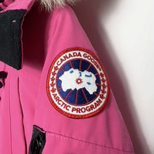 Rosa dunjacka från Canada Goose - Snygg rosa dunjacka från Canada Goose med stor huva och fluffig fuskpäls. Jackan har flera fickor, dragkedja och knappar framtill samt klassisk patch på ärmen. Perfekt för kalla dagar när du vill sticka ut med färg.