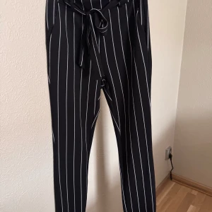 Svarta randiga byxor från SHEIN XS - Svarta byxor från SHEIN i storlek XS med vita vertikala ränder och knytband i midjan. Byxorna har en rak passform och är tillverkade i ett mjukt, syntetiskt material. Perfekta för en stilren och trendig look.