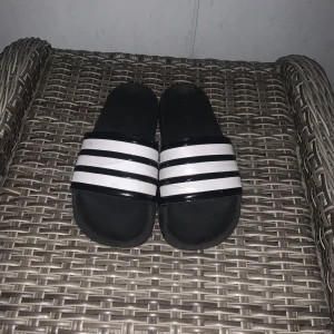 Svarta och vita Adidas tofflor - Klassiska Adidas sandaler i svart med tre vita ränder över foten och Adidas-logga på sidan. Snygg och enkel design med platt sula och öppen tå, perfekta för sommaren eller efter träningen. Tillverkade i syntetmaterial för en bekväm känsla.