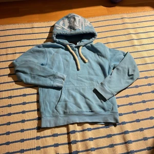 Blå hoodie från syna med tryck - Ljusblå hoodie från syna med fet graffiti-tryck på huvan och grova vita snören. Klassisk känguruficka framtill och mjukt material som känns skönt mot huden. Perfekt för dig som gillar streetstyle och vill sticka ut lite extra.