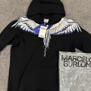 Marcelo Burlon svart hoodie med vingar - Svart hoodie från Marcelo Burlon med ikoniskt vingtryck i vitt och blått över axlar och bröst. Tröjan har huva med snörning och en stor magficka. Perfekt statement-plagg för dig som vill sticka ut.
