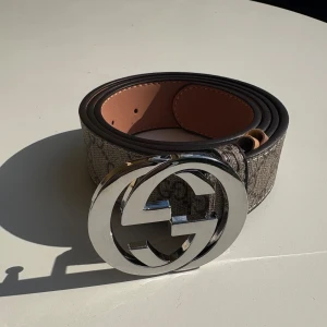 Gucci GG Supreme bälte med silverdetalj - Snyggt Gucci-bälte i klassiskt GG Supreme-mönster med bred rem i beige och brunt. Stor, rund spänne i blankt silver med dubbla G-loggan. Bältet är gjort i skinn och har en exklusiv känsla, perfekt för att lyfta vilken outfit som helst.