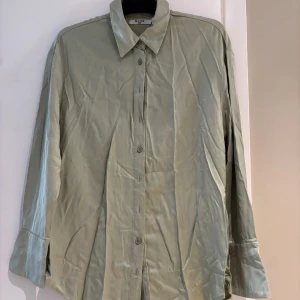 Ljusgrön skjorta från NA-KD, strl 34 - Snygg ljusgrön skjorta från NA-KD i storlek 34. Skjortan har klassisk krage, knäppning framtill och långa ärmar med breda manschetter. Materialet är mjukt och har en lätt glansig finish som ger en fräsch look.