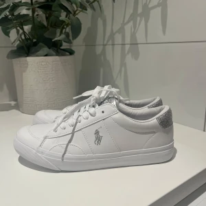Vita sneakers Polo Ralph Lauren glitter - Snygga vita sneakers från Polo Ralph Lauren med silverglittrande detaljer. Klassisk låg modell med snörning och logga på sidan. Helt nya och aldrig använda.