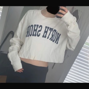 Vit croppad tröja John Galt - Vit croppad långärmad tröja från John Galt med blå texten 'NORTH SHORE' framtill. Tröjan är i ribbad bomull och har en relaxed, loose passform med långa ärmar. Perfekt för en chill och avslappnad stil.