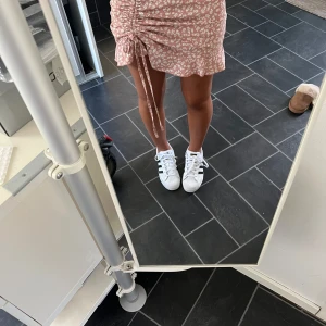 Rosa blommig kortkjol med snörning - Supersöt rosa kortkjol med vita blommor och volangkant. Kjolen har snörning på sidan som ger en snygg rynkad effekt och en feminin touch. Perfekt för varma dagar och lätt att matcha med sneakers eller sandaler.