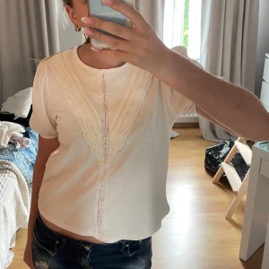 Vit blus med spetsdetaljer - Supersöt vit blus med korta puffärmar och rund halsringning. Framsidan har fina spetsdetaljer i V-form och längs mitten, vilket ger en romantisk och vintage-inspirerad vibe. Perfekt att matcha med jeans eller kjol för en chill och stilren look.